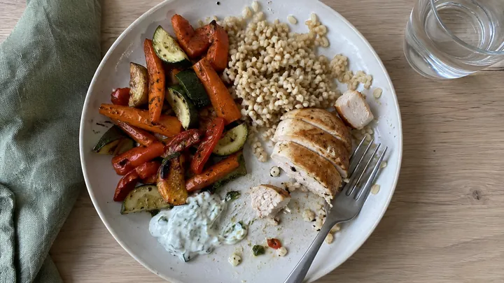 KI-generiertes Symbolbild: Teller von oben, halb gegessen: Ofengemüse aus Möhren, Zucchini und Paprika, eine Portion Graupen, Hähnchenbrust in Scheiben und Kräuterjoghurt, daneben ein Glas Wasser
