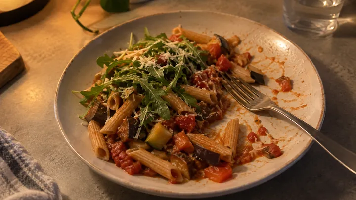KI-generiertes Symbolbild: Halb gegessener Teller Vollkornnudeln mit Tomaten-Gemüse-Soße, Rucola und Parmesan, eine Gabel liegt auf dem Teller, warmes Abendlicht
