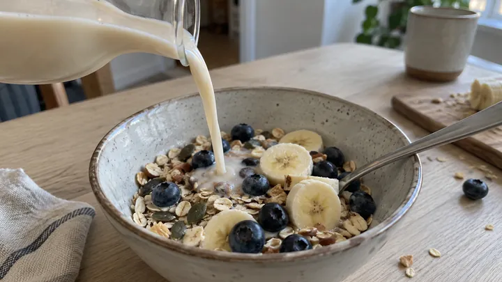 KI-generiertes Symbolbild: Hafermilch wird aus einer schlichten Glaskaraffe über eine Schüssel Müsli mit Heidelbeeren und Bananenscheiben gegossen