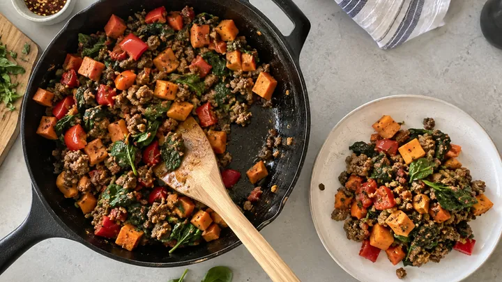 KI-generiertes Symbolbild: Gusseiserne Pfanne von oben mit angebratenem Rinderhack, Süßkartoffelwürfeln, Paprika und Spinat, ein Holzwender steckt darin, daneben ein Teller mit einer Portion